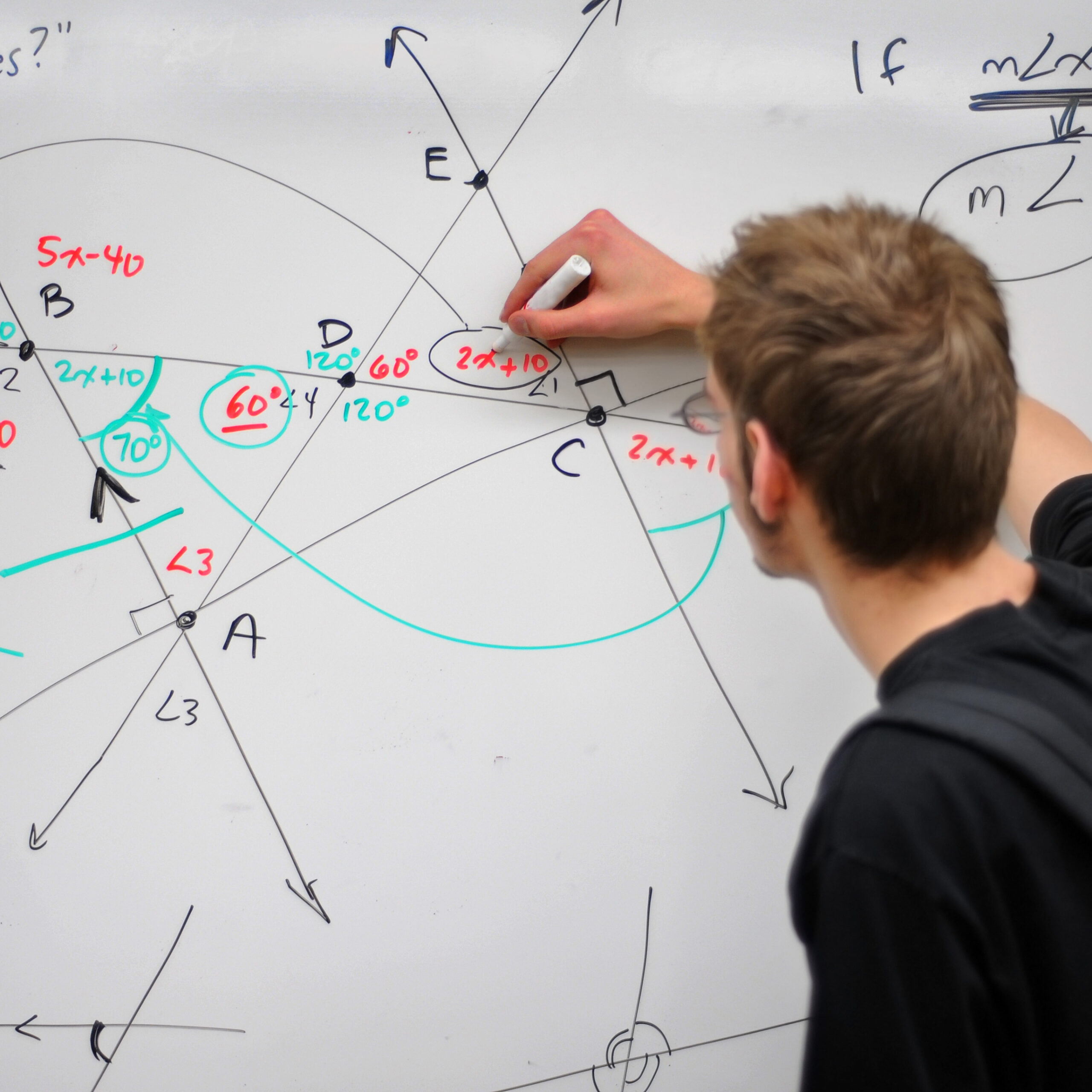 A young man stands at a whiteboard filled with a hand-drawn geometric diagram—intersecting lines, arcs, and labeled points (A, B, C, D, E)—annotated with colored angle measurements (e.g. 60°, 70°) and algebraic expressions like “2x+10” and “5x–40.” He’s holding a whiteboard marker and appears to be explaining or solving the geometry problem.